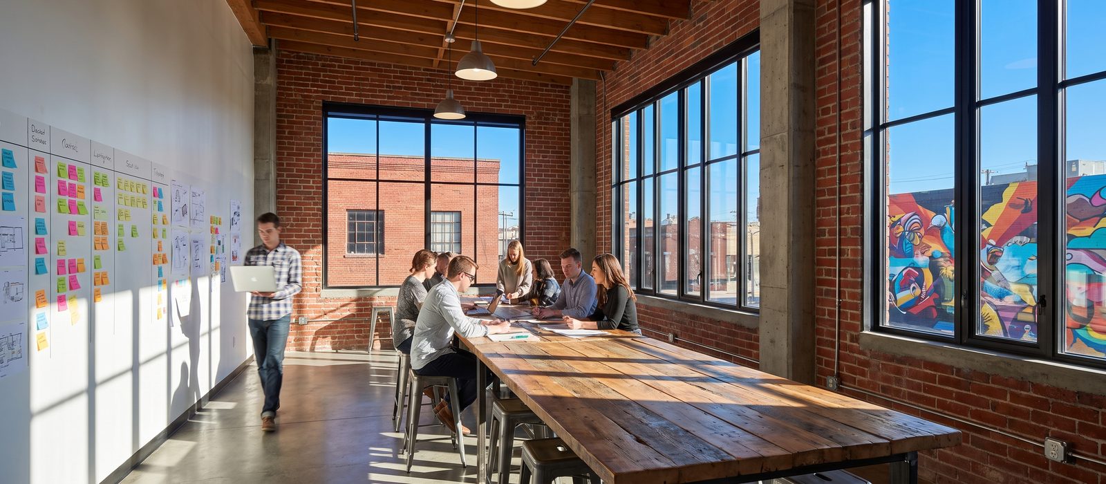 A mixed young design team in a bright converted-warehouse studio in RiNo: one walking through with a laptop, others at a wall of sticky notes and along a long timber table; hard sunlight, red brick and a mural across the street through the tall windows.