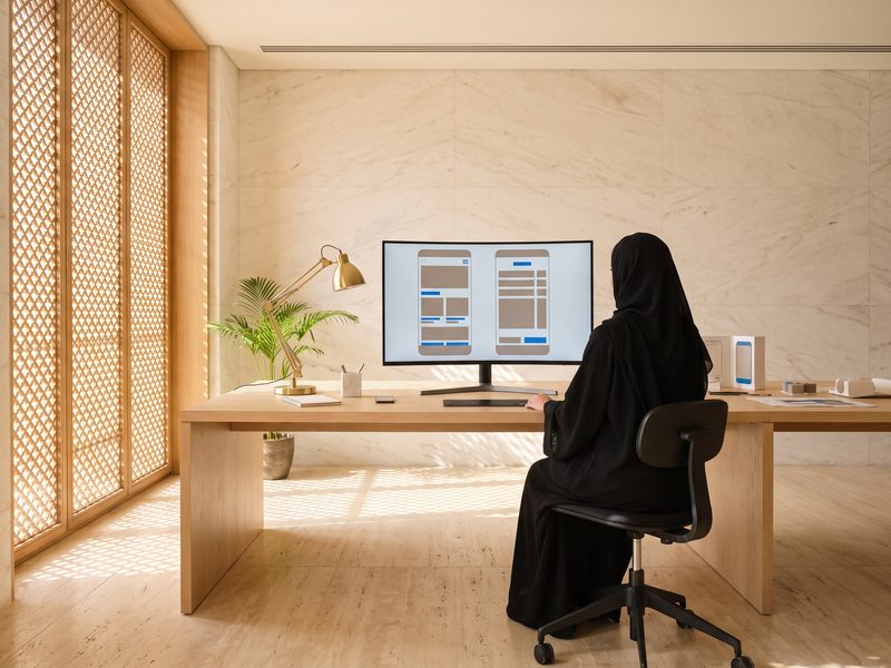 An Emirati designer in an abaya at a monitor showing the same app interface mirrored right-to-left and left-to-right as grey blocks, warm light through a mashrabiya screen.