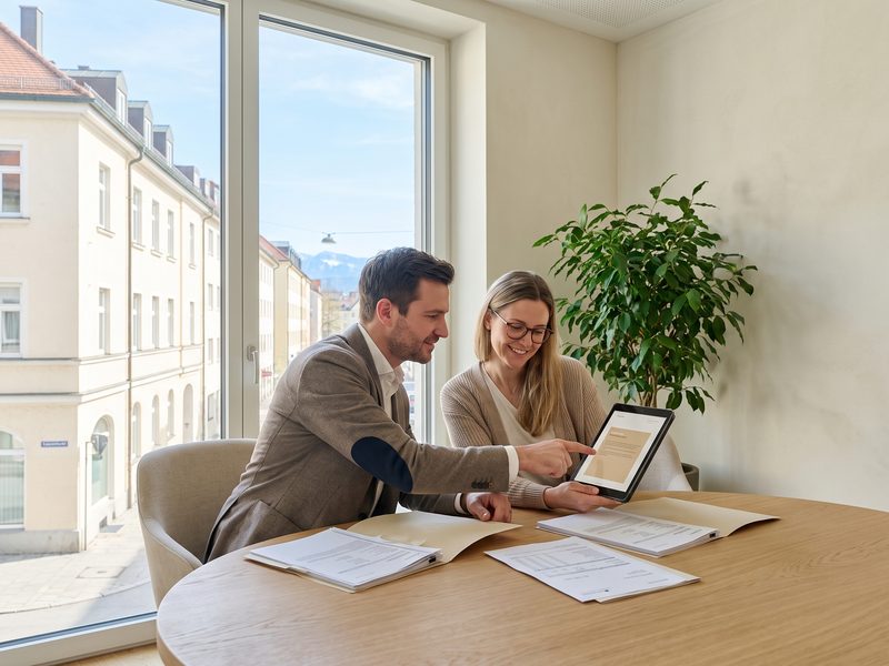 An advisor and an older client with reading glasses reviewing a document on a tablet beside printed contracts at an oak desk by a bright window.