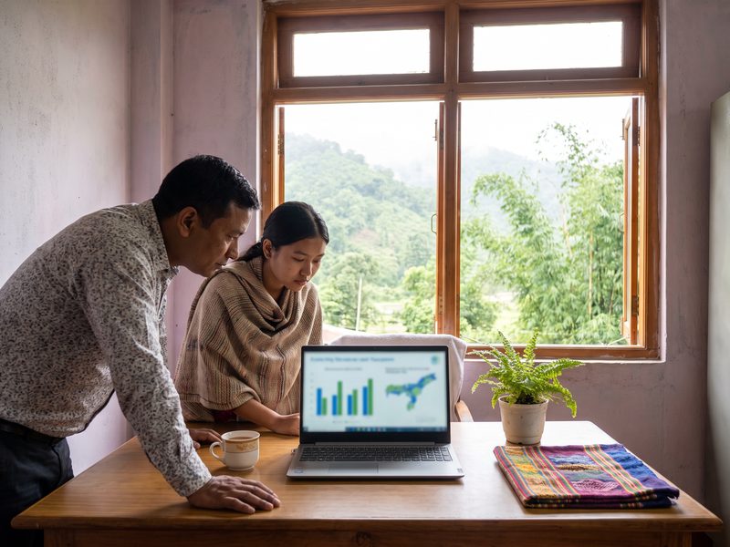 A district officer and a colleague looking together at a laptop dashboard at a bright window desk in a whitewashed Guwahati office, green hills and mist outside.